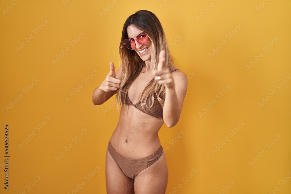 Young hispanic woman wearing bikini over yellow background pointing fingers to camera with happy and funny face. good energy and vibes. Young hispanic woman wearing bikini over yellow background pointing fingers to camera with happy and funny face. good energy and vibes.