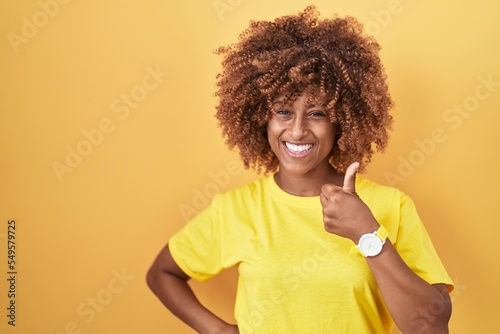 Young hispanic woman with curly hair standing over yellow background doing happy thumbs up gesture with hand. approving expression looking at the camera showing success.