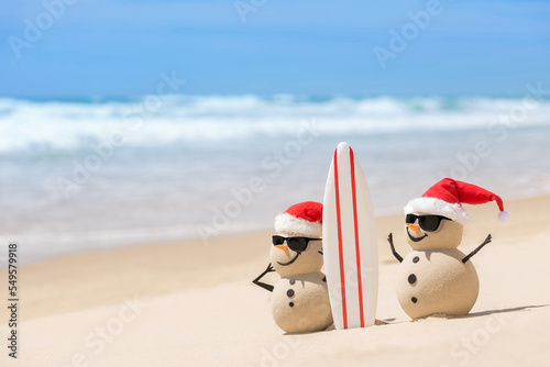 Two Sandy Christmas Snowmen are watching the waves, standing on beautiful beach with a surf board, wearing sunnies