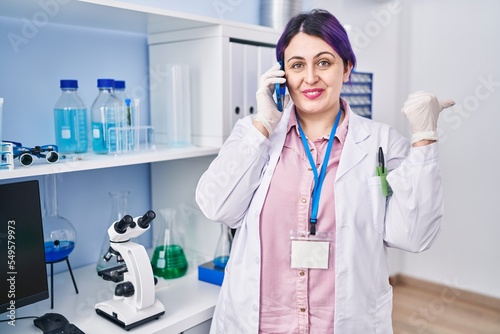 Plus size woman wit purple hair working at scientist laboratory speaking on g the phone pointing thumb up to the side smiling happy with open mouth