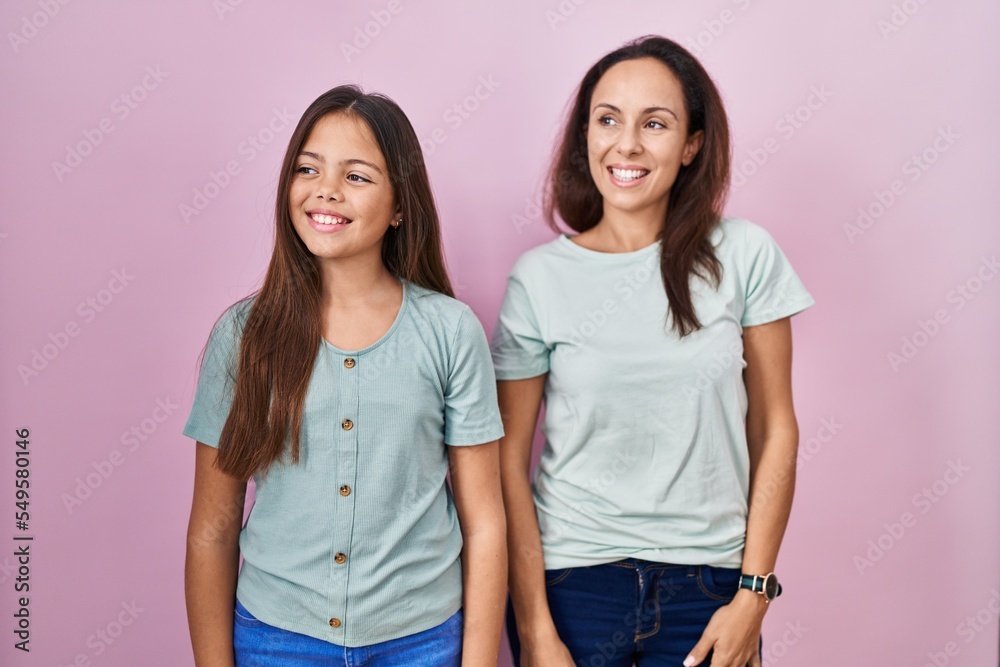 Young mother and daughter standing over pink background looking away to side with smile on face, natural expression. laughing confident. Young mother and daughter standing over pink background looking away to side with smile on face, natural expression. laughing confident.
