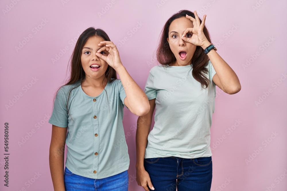 Young mother and daughter standing over pink background doing ok gesture shocked with surprised face, eye looking through fingers. unbelieving expression. Young mother and daughter standing over pink background doing ok gesture shocked with surprised face, eye looking through fingers. unbelieving expression.