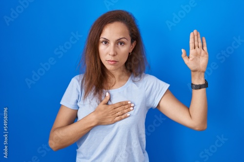 Brunette woman standing over blue background swearing with hand on chest and open palm  making a loyalty promise oath