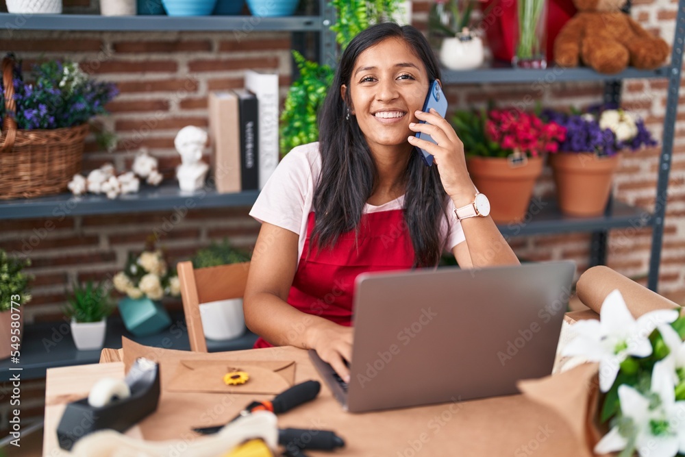 Young beautiful hispanic woman florist talking on smartphone using laptop at flower shop Young beautiful hispanic woman florist talking on smartphone using laptop at flower shop