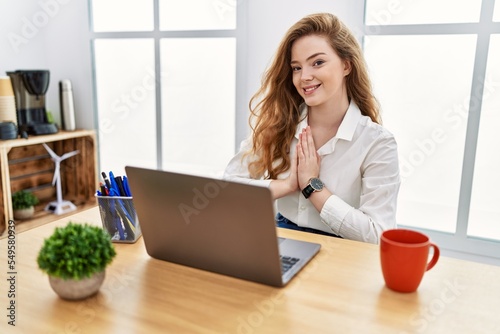 Young caucasian woman working at the office using computer laptop praying with hands together asking for forgiveness smiling confident.