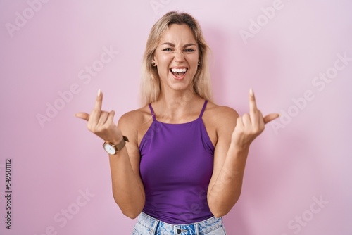 Young blonde woman standing over pink background showing middle finger doing fuck you bad expression, provocation and rude attitude. screaming excited