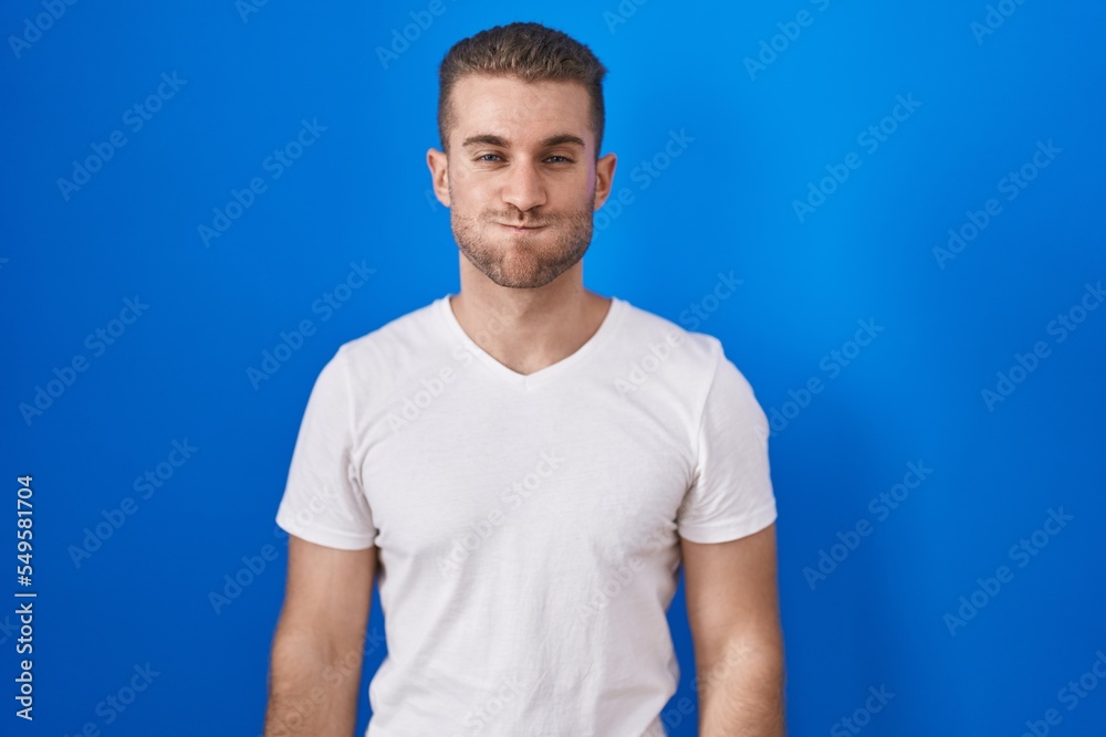 Young caucasian man standing over blue background puffing cheeks with funny face. mouth inflated with air, crazy expression. Young caucasian man standing over blue background puffing cheeks with funny face. mouth inflated with air, crazy expression.