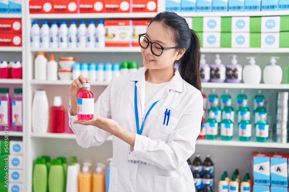 Young chinese woman pharmacist smiling confident holding medication bottle at pharmacy