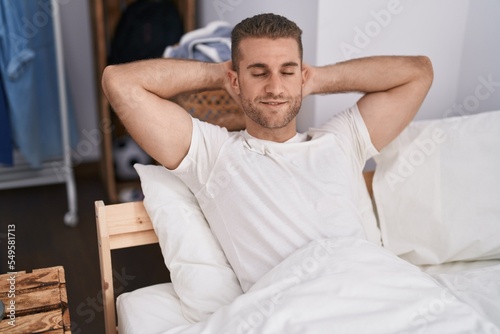 Young caucasian man smiling confident relaxed with hands on bed at bedroom