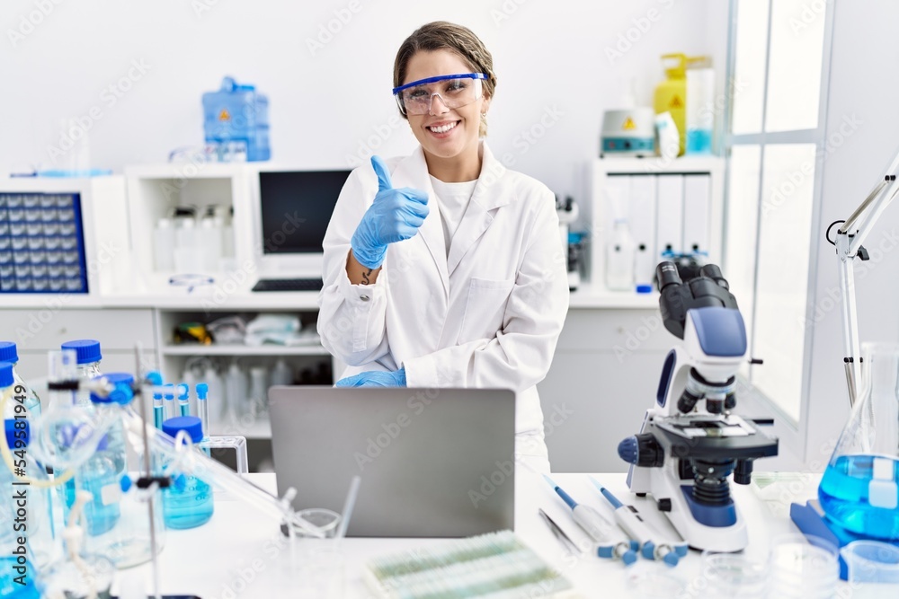 Young hispanic woman wearing scientist uniform working at laboratory smiling happy and positive, thumb up doing excellent and approval sign Young hispanic woman wearing scientist uniform working at laboratory smiling happy and positive, thumb up doing excellent and approval sign