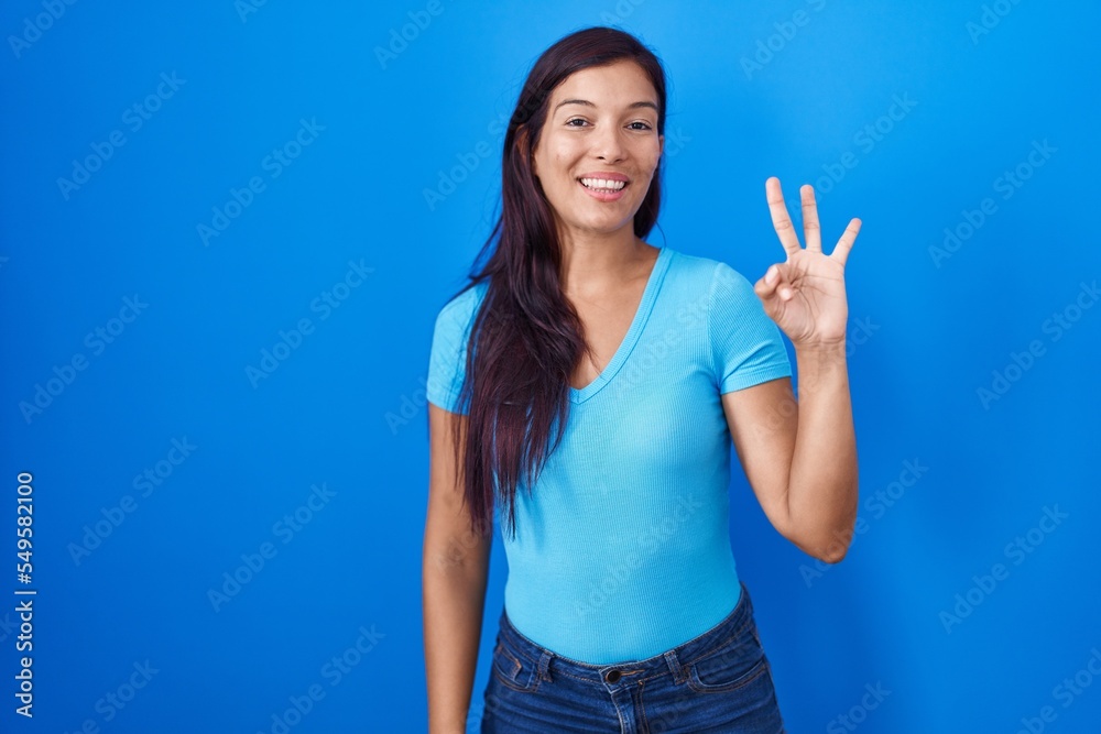 Fototapeta premium Young hispanic woman standing over blue background showing and pointing up with fingers number three while smiling confident and happy.
