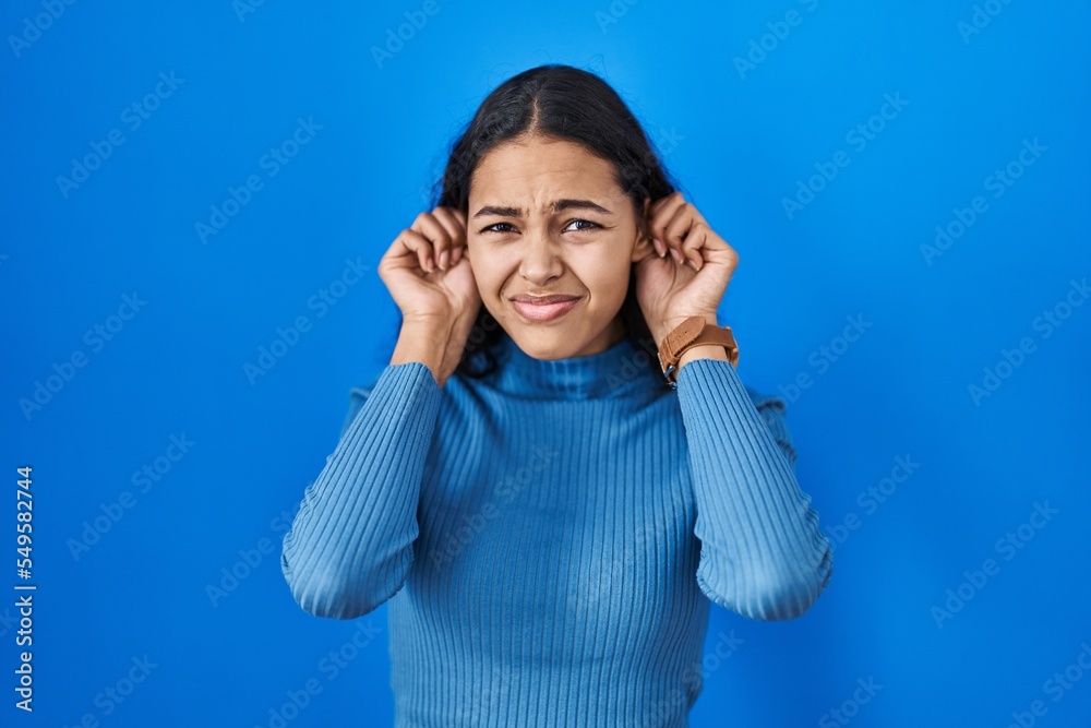 Young brazilian woman standing over blue isolated background covering ears with fingers with annoyed expression for the noise of loud music. deaf concept. Young brazilian woman standing over blue isolated background covering ears with fingers with annoyed expression for the noise of loud music. deaf concept.