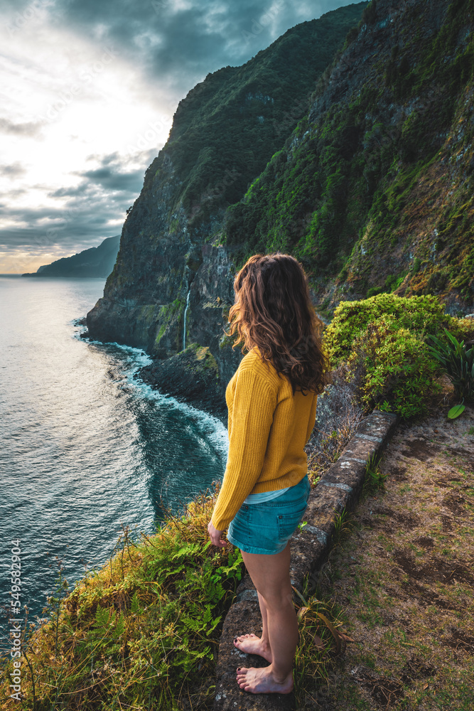 Naklejka premium Sporty woman looking at waterfall flowing into the sea in atmospheric morning atmosphere. Viewpoint Véu da Noiva, Madeira Island, Portugal, Europe.