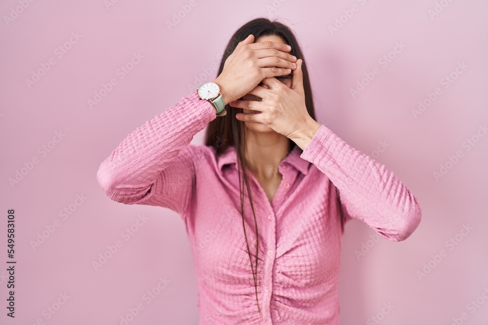 Young hispanic woman standing over pink background covering eyes and mouth with hands, surprised and shocked. hiding emotion