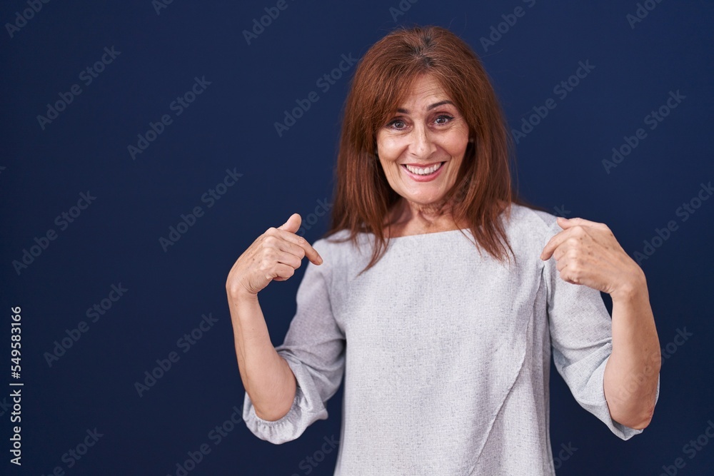 Fototapeta premium Middle age woman standing over blue background looking confident with smile on face, pointing oneself with fingers proud and happy.