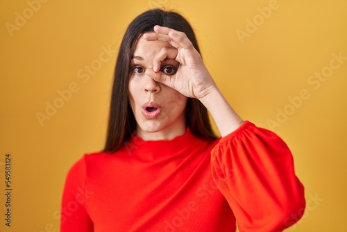 Young hispanic woman standing over yellow background doing ok gesture shocked with surprised face, eye looking through fingers. unbelieving expression. © Krakenimages.com