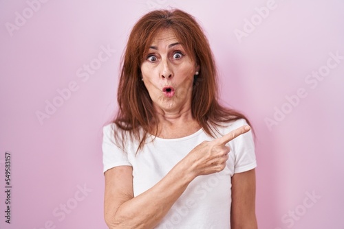 Middle age woman standing over pink background surprised pointing with finger to the side, open mouth amazed expression.