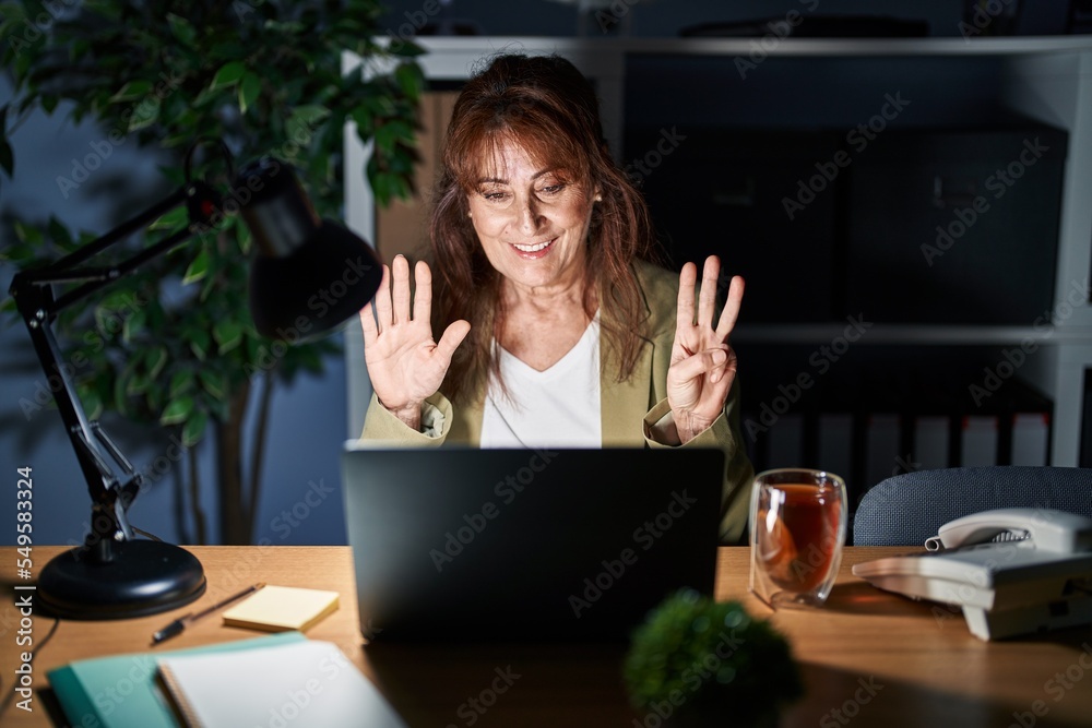 Middle age hispanic woman working using computer laptop at night showing and pointing up with fingers number eight while smiling confident and happy. Middle age hispanic woman working using computer laptop at night showing and pointing up with fingers number eight while smiling confident and happy.