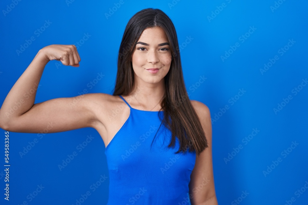 Hispanic woman standing over blue background strong person showing arm muscle, confident and proud of power
