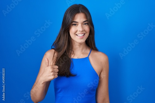 Hispanic woman standing over blue background doing happy thumbs up gesture with hand. approving expression looking at the camera showing success.