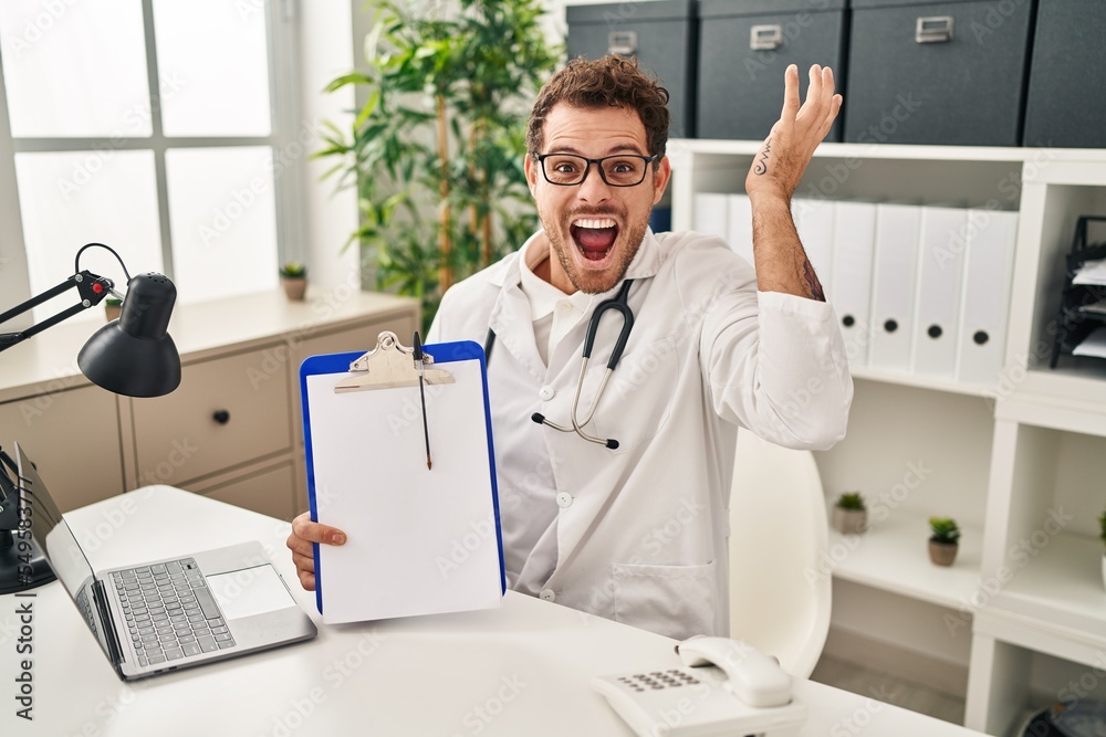 Young hispanic man wearing doctor stethoscope holding clipboard celebrating victory with happy smile and winner expression with raised hands