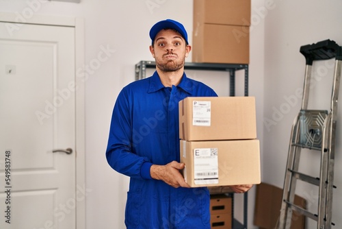 Young hispanic man working on moving service holding boxes puffing cheeks with funny face. mouth inflated with air, catching air. © Krakenimages.com