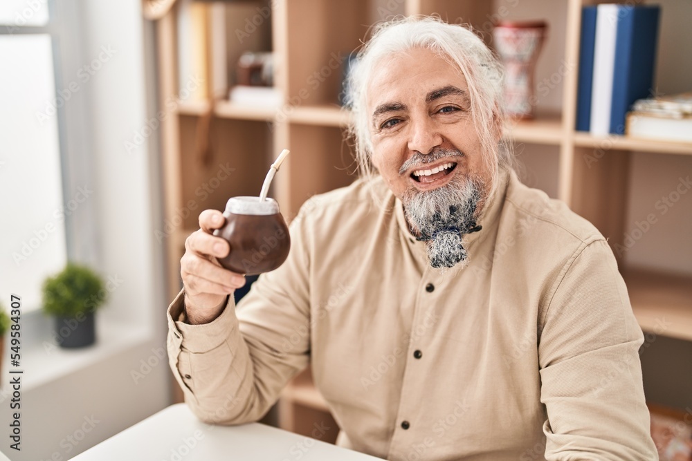 Middle age man with grey hair drinking mate infusion looking positive and happy standing and smiling with a confident smile showing teeth Middle age man with grey hair drinking mate infusion looking positive and happy standing and smiling with a confident smile showing teeth