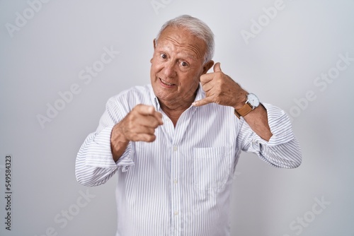 Senior man with grey hair standing over isolated background smiling doing talking on the telephone gesture and pointing to you. call me.
