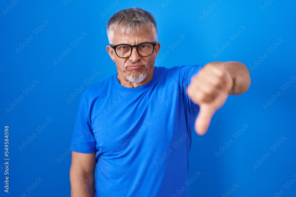 Hispanic man with grey hair standing over blue background looking unhappy and angry showing rejection and negative with thumbs down gesture. bad expression. Hispanic man with grey hair standing over blue background looking unhappy and angry showing rejection and negative with thumbs down gesture. bad expression.