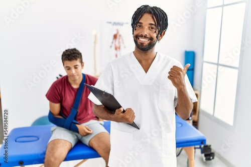 Young hispanic man working at pain recovery clinic with a man with broken arm pointing to the back behind with hand and thumbs up, smiling confident