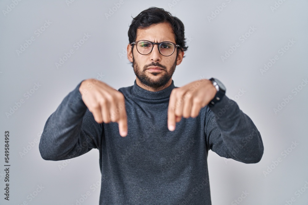 Handsome latin man standing over isolated background pointing down looking sad and upset, indicating direction with fingers, unhappy and depressed. Handsome latin man standing over isolated background pointing down looking sad and upset, indicating direction with fingers, unhappy and depressed.