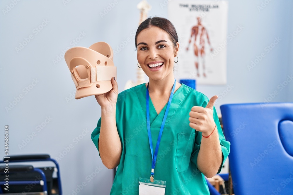 Young hispanic woman holding cervical neck collar at the clinic smiling happy and positive, thumb up doing excellent and approval sign Young hispanic woman holding cervical neck collar at the clinic smiling happy and positive, thumb up doing excellent and approval sign