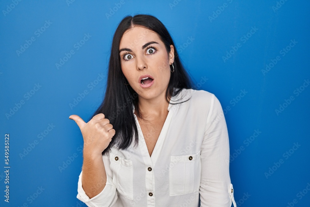 Young hispanic woman standing over blue background surprised pointing with hand finger to the side, open mouth amazed expression.