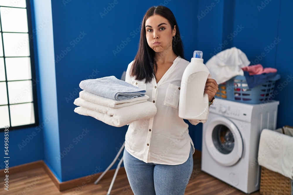 Young hispanic woman holding clean laundry and detergent bottle puffing cheeks with funny face. mouth inflated with air, catching air. - obrazy, fototapety, plakaty 