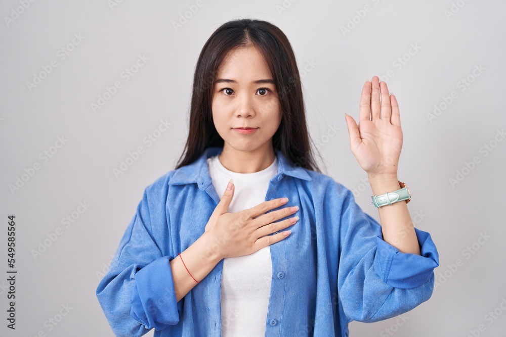 Young chinese woman standing over white background swearing with hand on chest and open palm, making a loyalty promise oath Young chinese woman standing over white background swearing with hand on chest and open palm, making a loyalty promise oath