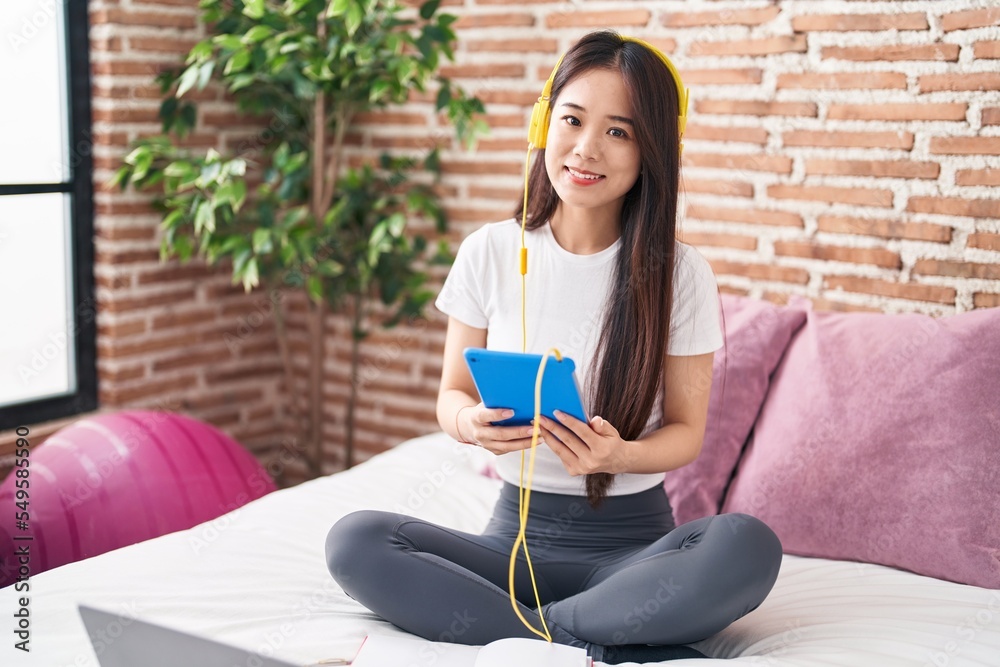 Young chinese woman using touchpad and headphones sitting on bed at bedroom Young chinese woman using touchpad and headphones sitting on bed at bedroom
