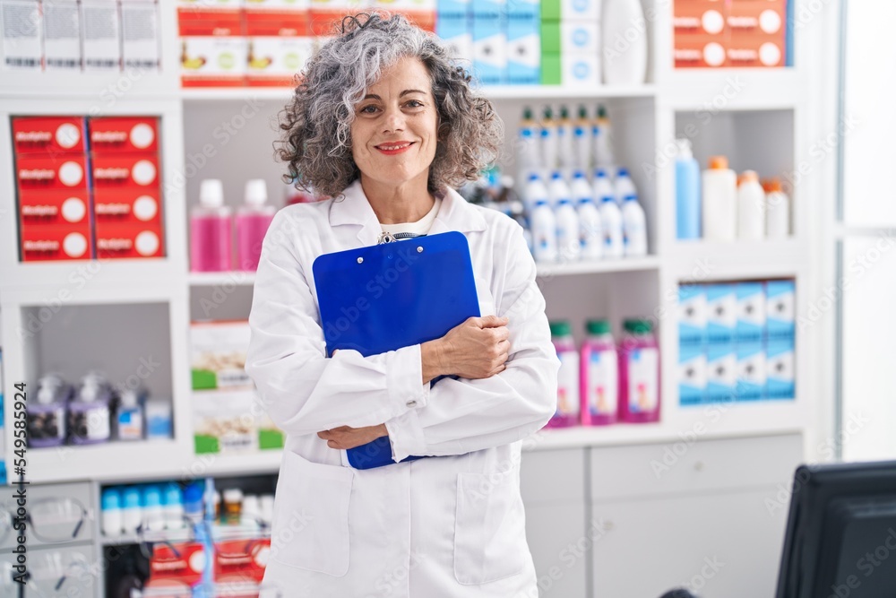 Middle age grey-haired woman pharmacist holding clipboard at pharmacy