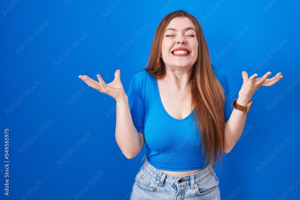 Redhead woman standing over blue background celebrating mad and crazy for success with arms raised and closed eyes screaming excited. winner concept Redhead woman standing over blue background celebrating mad and crazy for success with arms raised and closed eyes screaming excited. winner concept