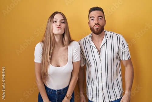 Young couple standing over yellow background looking at the camera blowing a kiss on air being lovely and sexy. love expression.