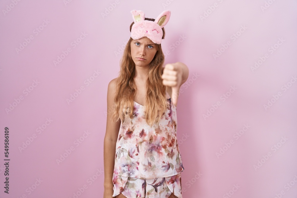 Young caucasian woman wearing sleep mask and pajama looking unhappy and angry showing rejection and negative with thumbs down gesture. bad expression.