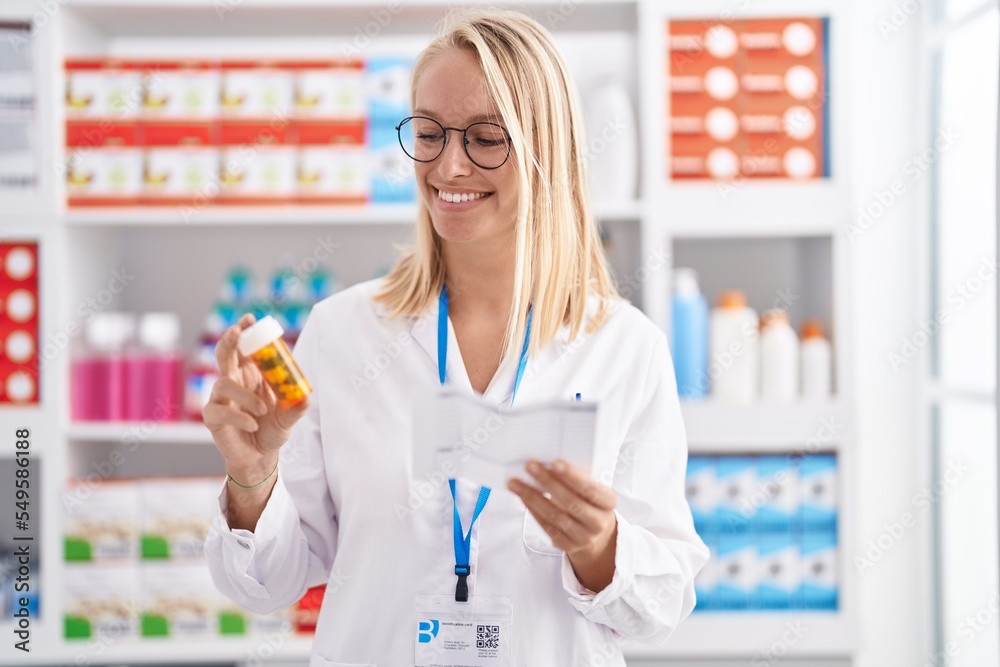 Young blonde woman pharmacist holding pills bottle reading prescription at pharmacy