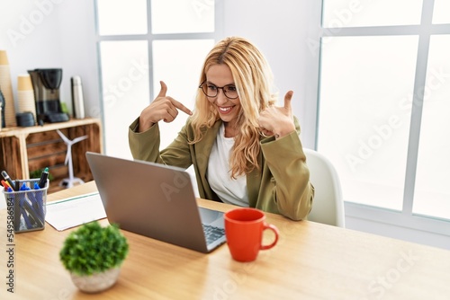 Beautiful blonde woman working at the office with laptop looking confident with smile on face, pointing oneself with fingers proud and happy.