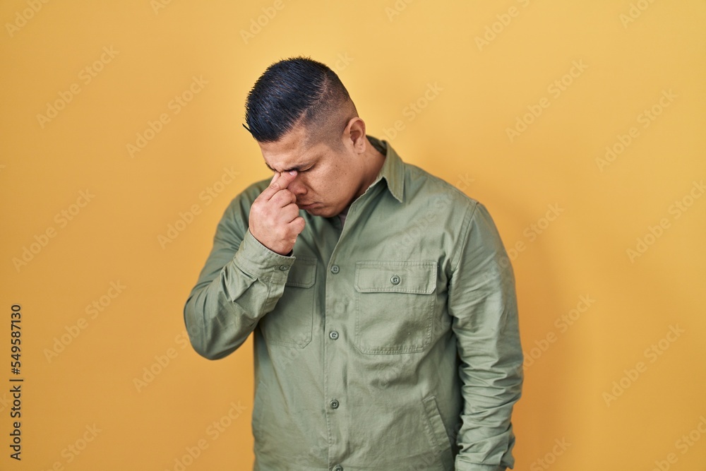 Hispanic young man standing over yellow background tired rubbing nose and eyes feeling fatigue and headache. stress and frustration concept. Hispanic young man standing over yellow background tired rubbing nose and eyes feeling fatigue and headache. stress and frustration concept.