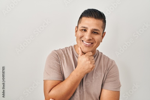 Hispanic young man standing over white background looking confident at the camera smiling with crossed arms and hand raised on chin. thinking positive.