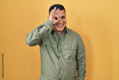 Hispanic young man standing over yellow background doing ok gesture with hand smiling  eye looking through fingers with happy face.
