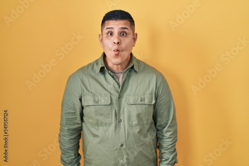 Hispanic young man standing over yellow background making fish face with lips, crazy and comical gesture. funny expression.