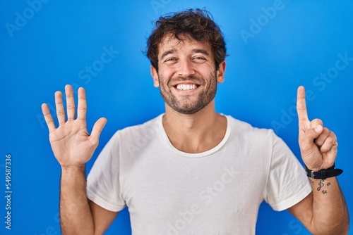 Hispanic young man standing over blue background showing and pointing up with fingers number six while smiling confident and happy.