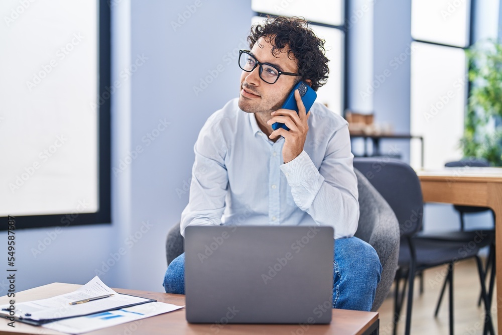 Young hispanic man business worker using laptop talking on smartphone at office Young hispanic man business worker using laptop talking on smartphone at office
