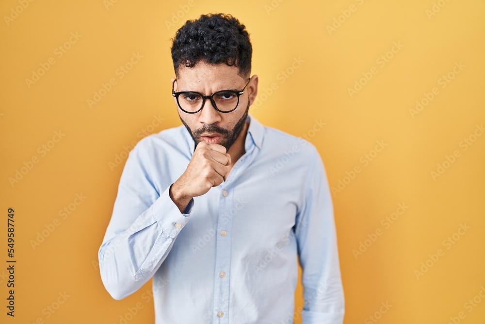 Hispanic man with beard standing over yellow background feeling unwell and coughing as symptom for cold or bronchitis. health care concept. Hispanic man with beard standing over yellow background feeling unwell and coughing as symptom for cold or bronchitis. health care concept.