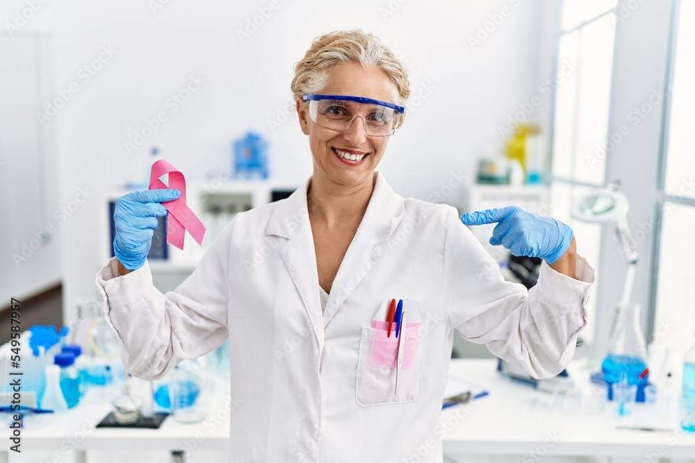 Middle age blonde woman working at laboratory looking for breast cancer cure pointing finger to one self smiling happy and proud Middle age blonde woman working at laboratory looking for breast cancer cure pointing finger to one self smiling happy and proud